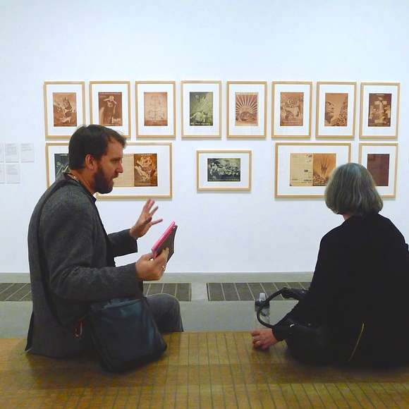 Man and woman seated in discussion at Tate Modern in front of a wall of framed sepia-tone prints.