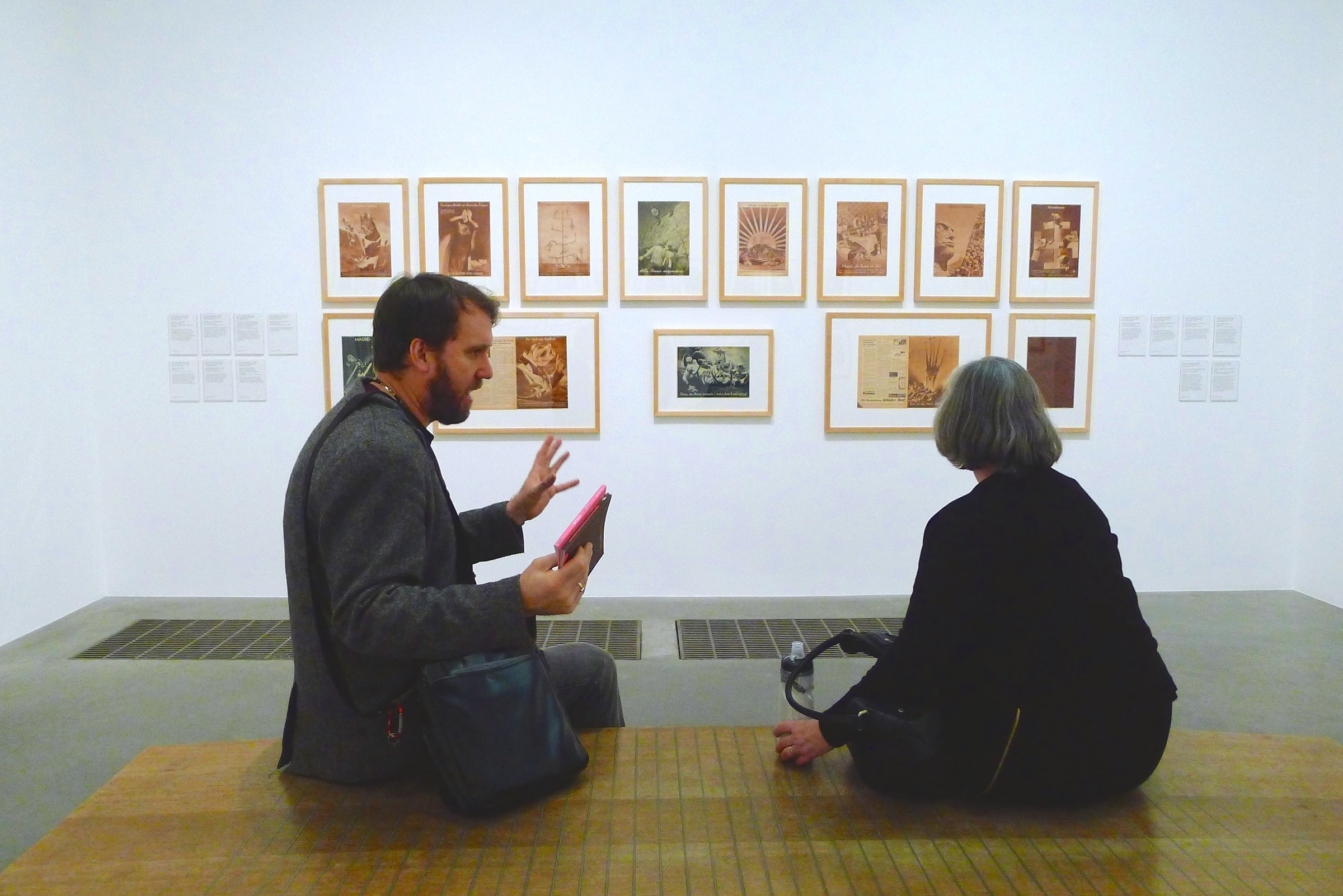 Man and woman seated in discussion at Tate Modern in front of a wall of framed sepia-tone prints.